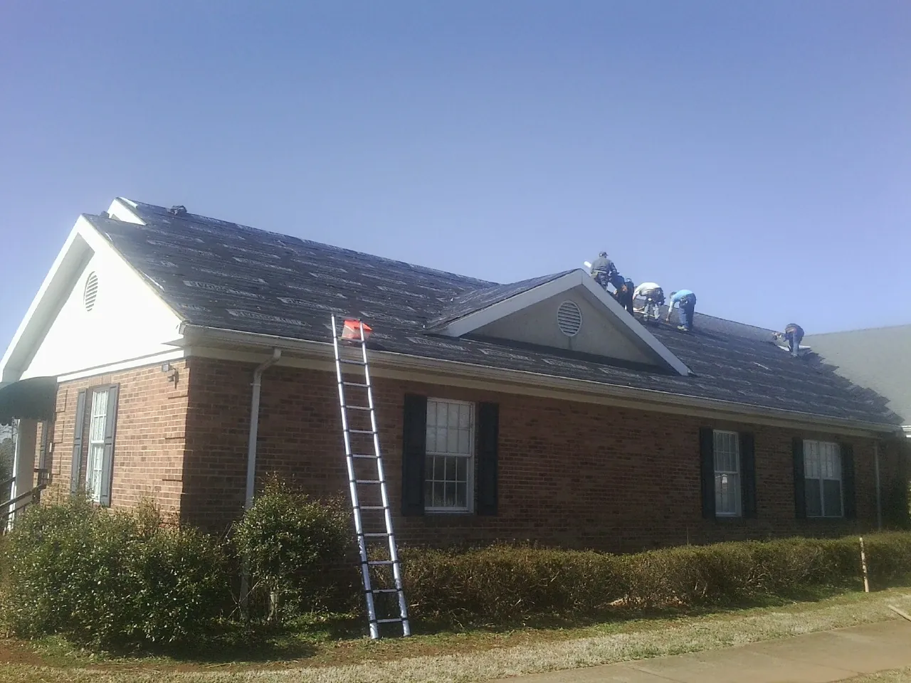 Crew tearing shingles off a brick ranch roof with a ladder set — Emergency Roof Repair work by our crew in New Franken