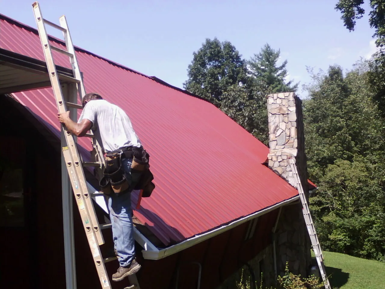 Roofer on a ladder beside a red metal roof and stone chimney — Soffit & Fascia Repair by Roofing Brothers in New Franken